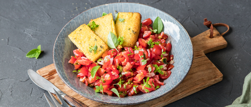 Grauer Teller mit Polentaschnitten und frischem Tomatensalat mit Kräutern auf einem Holzbrett, daneben Besteck.