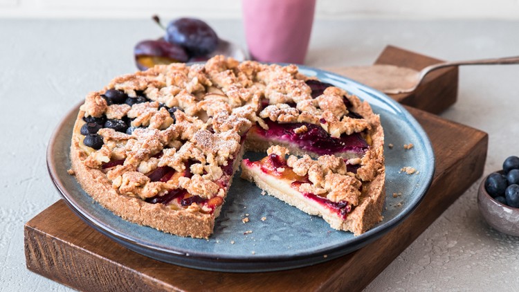 Runder Streuselkuchen mit Zwetschgen und Blaubeeren auf blauem Teller, daneben rosa Becher und kleine Schale mit Blaubeeren.