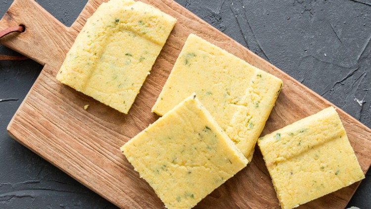 Vier quadratische Stücke Maisgrießbrot mit Kräutern liegen auf einem hölzernen Schneidebrett mit Lederschlaufe.