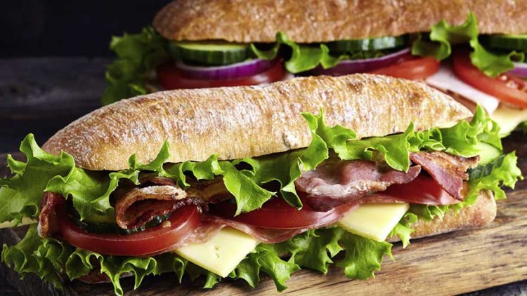 Zwei belegte Baguettes mit Salat, Tomaten, Gurken, roten Zwiebeln, Käse und knusprigem Bacon auf einem Holzbrett.