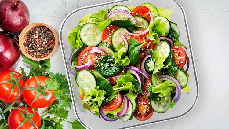 Ein frischer Gemüsesalat mit Gurken, Tomaten, roten Zwiebeln, Blattsalat und Spinat in einer Glasschüssel, daneben Tomaten und Gewürze.