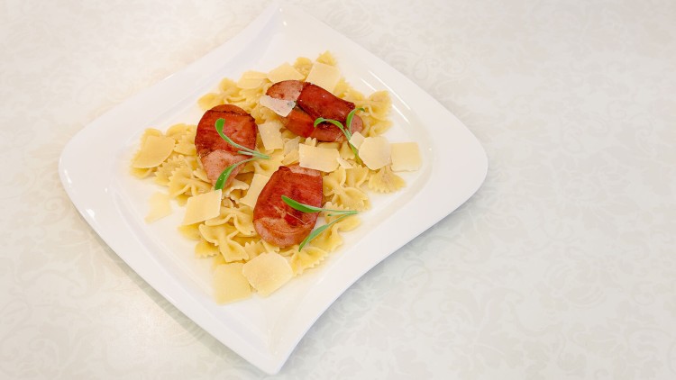 Farfalle-Nudeln mit gebratener Wurst, grüner Garnitur und Käsestücken auf einem weißen Teller.