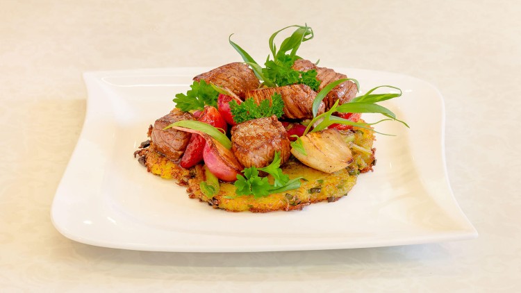 Gebratene Fleischstücke mit frischen Kräutern, Gemüse und Rösti auf einem weißen, quadratisch geschwungenen Teller.
