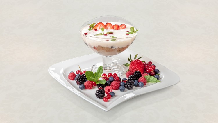 Ein Dessertglas mit Schichtdessert, garniert mit Erdbeeren und Minze, steht auf einem Teller mit gemischten Beeren.