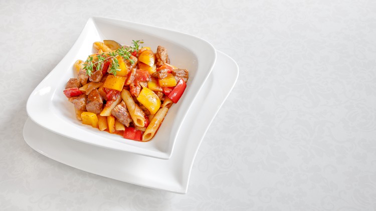 Weiße Schale mit Penne, roter und gelber Paprika, Fleischstücken in Tomatensoße, garniert mit Thymianzweigen auf hellem Untergrund.