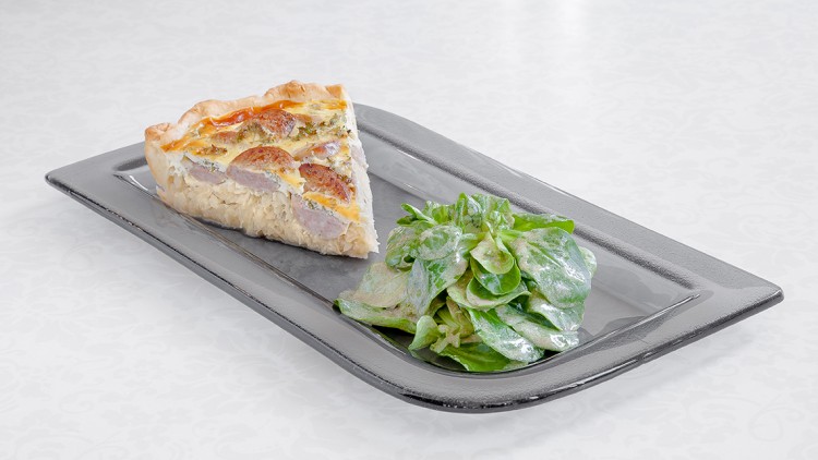 Ein Stück Quiche mit Wurst und Kraut liegt auf einem rechteckigen Teller neben einem kleinen Salat mit heller Soße.