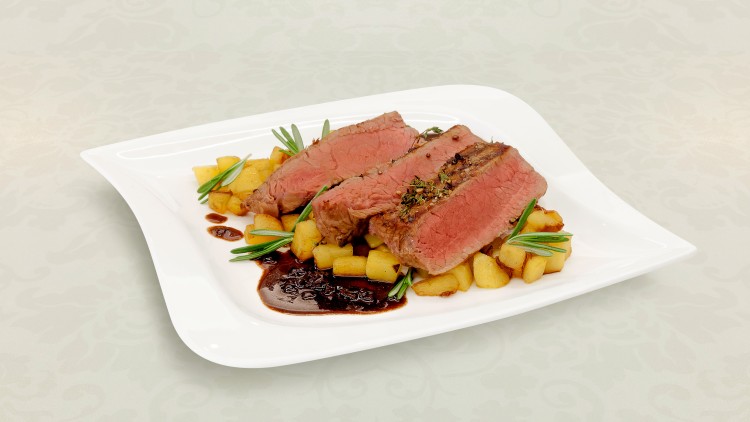 Drei Stücke rosa gebratenes Fleisch mit Kräutern auf Kartoffelwürfeln und dunkler Sauce auf weißem Teller mit Verzierung.