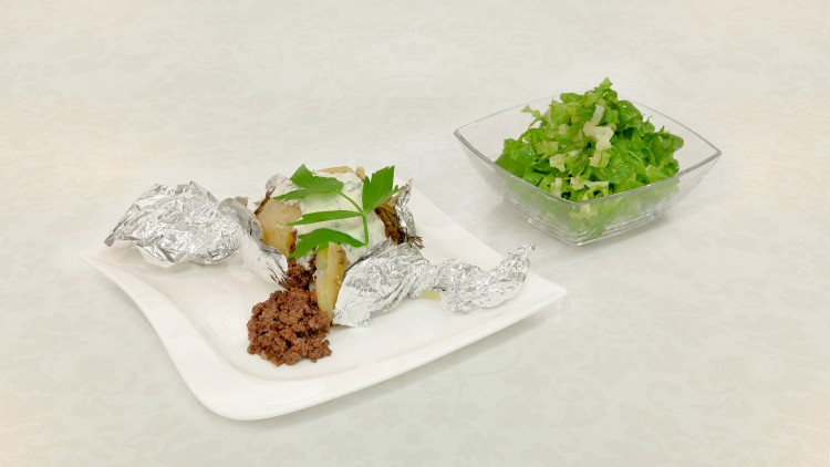 Auf einem weißen Teller liegen zwei Folienkartoffeln mit Kräutern und Hackfleisch, daneben steht eine Glasschüssel mit grünem Salat.