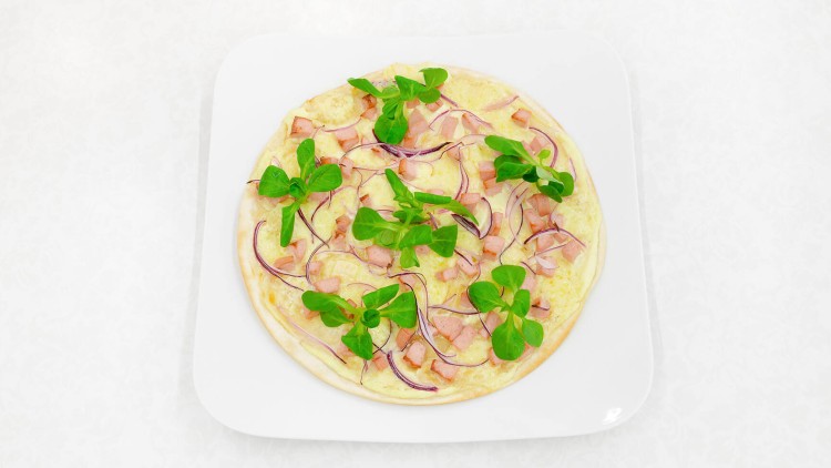 Flammkuchen mit gehacktem Schinken, roten Zwiebelstreifen und Salatblättern auf einem weißen quadratischen Teller.