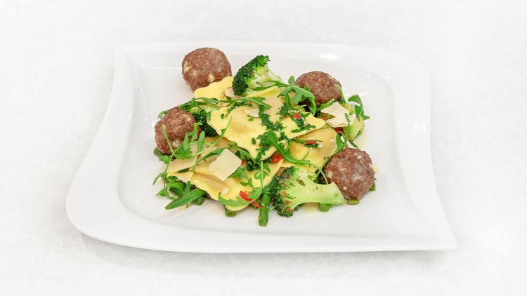 Ein Teller mit Ravioli, Brokkoli, Fleischbällchen, Rucola, Chilistreifen und Parmesanhobeln auf weißem Untergrund.