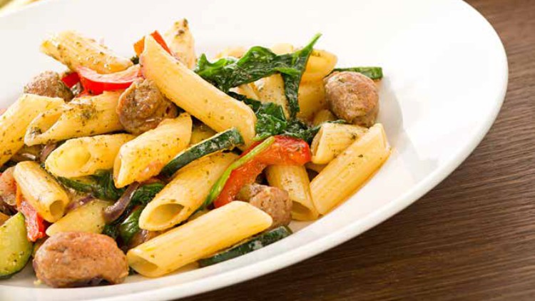 Zwei weiße Teller mit Penne-Nudeln, Gemüse und kleinen Fleischbällchen, im Hintergrund ein Glas mit grüner Pesto-Soße.