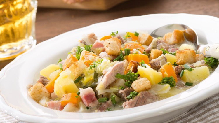Ein Teller mit Gemüsesuppe, Croutons, Kartoffeln und Fleischstücken, daneben Bier, Brot und ein Salat im Hintergrund.