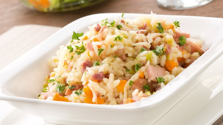 Weiße Schale mit Reisgericht aus Gemüse, Speck und Kräutern, dahinter ein Salat in einer Glasschüssel und ein Glas Weißwein.