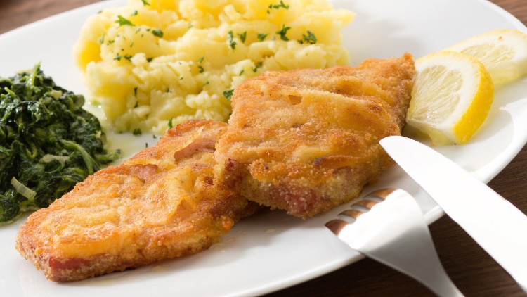 Panierte Schnitzel mit Zitronenscheiben, Kartoffelpüree mit Petersilie und Spinat auf einem Teller, daneben Besteck.