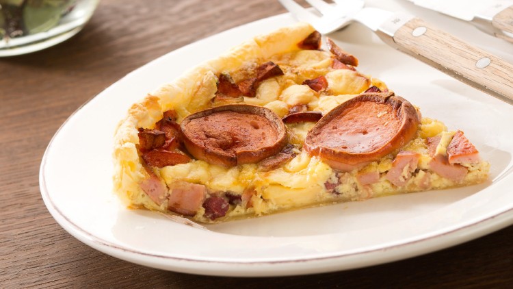 Ein Stück herzhafte Quiche mit Wurst und Schinken auf einem weißen Teller neben Messer und Gabel, im Hintergrund Salat und Serviette.