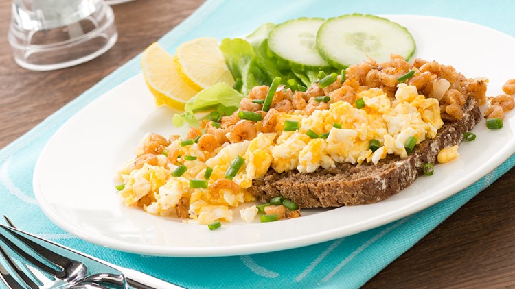 Ein Teller mit Rührei, Krabben und Schnittlauch auf Brot, dekoriert mit Salat, Gurkenscheiben und Zitrone, daneben Besteck.