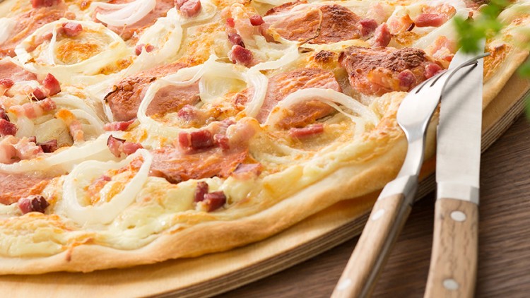 Flammkuchen mit Zwiebeln und Speck auf einem Holzbrett, daneben Messer und Gabel, im Hintergrund Kräuter und ein Glas Weißwein.
