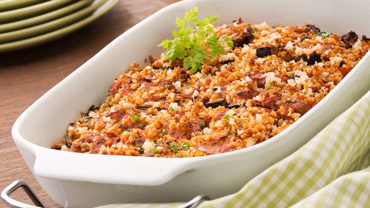 Eine weiße Auflaufform mit braun überbackenem Crumble und grünem Kraut darauf, daneben ein kariertes Tuch und Ölflasche.
