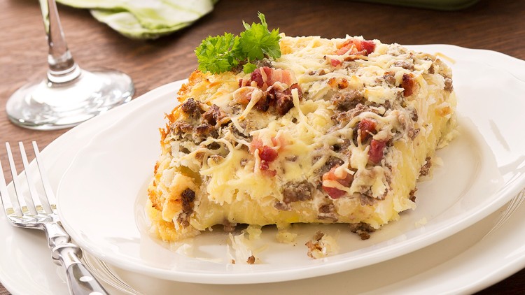 Ein Stück Auflauf mit Hackfleisch, Käse und Kräuterdekoration auf weißem Teller neben grünem Textiltuch und Auflaufform.