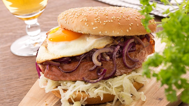 Burger mit Spiegelei, Zwiebeln und Krautsalat auf Holzbrett, daneben ein Glas Bier, Besteck und Petersilie im Vordergrund.