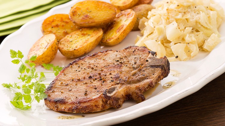 Gegrilltes Kotelett, goldbraune Kartoffeln und Krautsalat auf einem weißen Teller mit Kräuterdekoration, Bierkrug im Hintergrund.