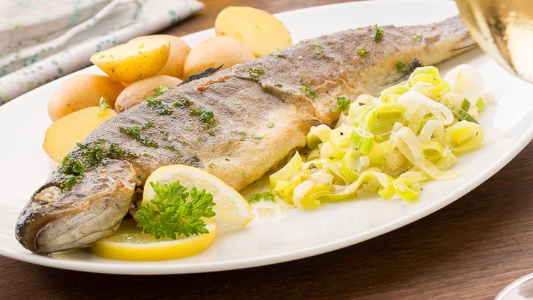 Gegrillte Forelle mit Zitrone, Petersilie, gekochten Kartoffeln und Lauchstreifen auf einem weißen Teller, daneben ein Weinglas.