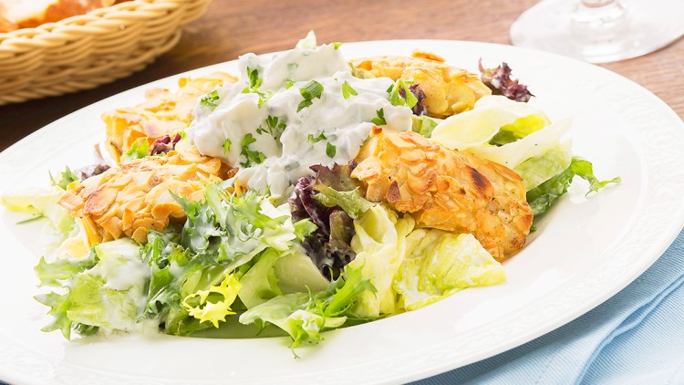 Teller mit gemischtem Salat, panierten Hähnchenstücken und Kräuterdip, daneben ein Weinglas und ein Brotkörbchen.