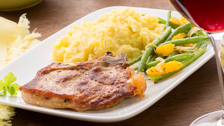 Ein Teller mit gebratenem Fleisch, Kartoffelpüree und grünen Bohnen mit Orangenscheiben, daneben ein Glas Rotwein.