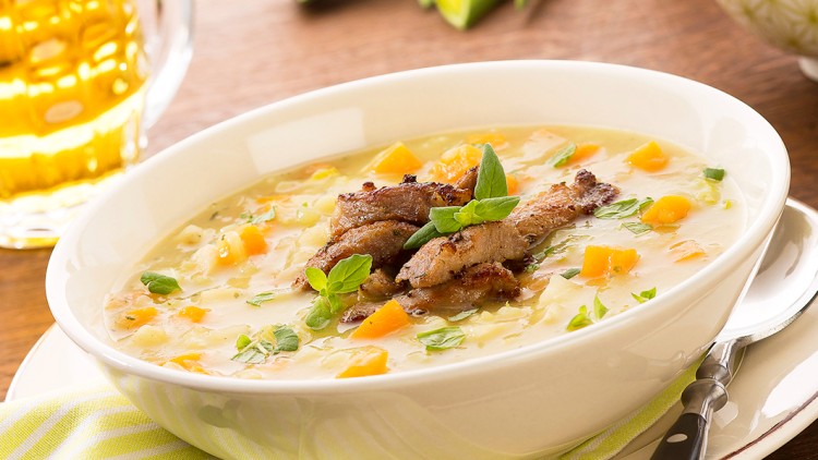 Eine weiße Schale mit Gemüsesuppe, Fleischstücken und Kräutern steht auf einem gestreiften grünen Tuch mit Löffel.