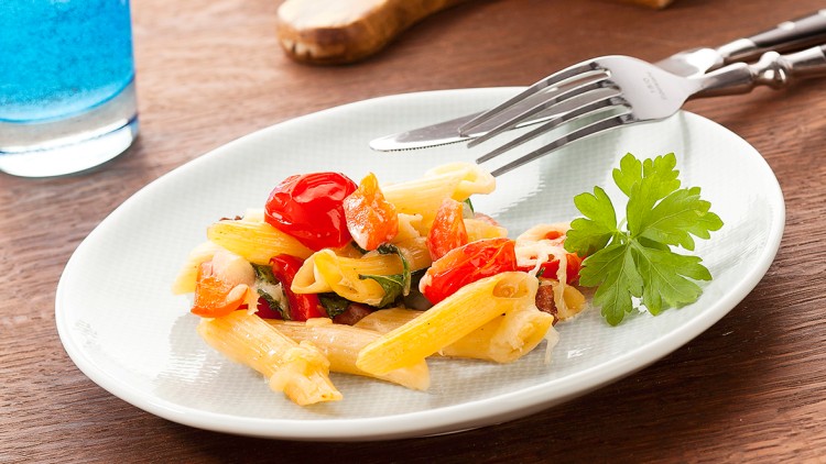 Ein Teller mit Penne, Tomaten, Kräutern und Petersilie neben Messer, Gabel, Glas Wasser und einer Auflaufform auf Holzuntergrund.