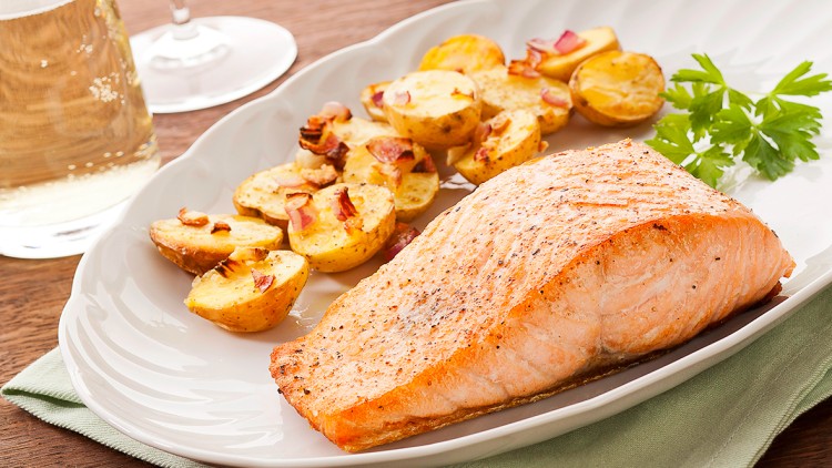 Gebratenes Lachsfilet mit ofengebackenen Kartoffelscheiben und Petersilie auf weißem Teller, daneben ein Glas Weißwein.