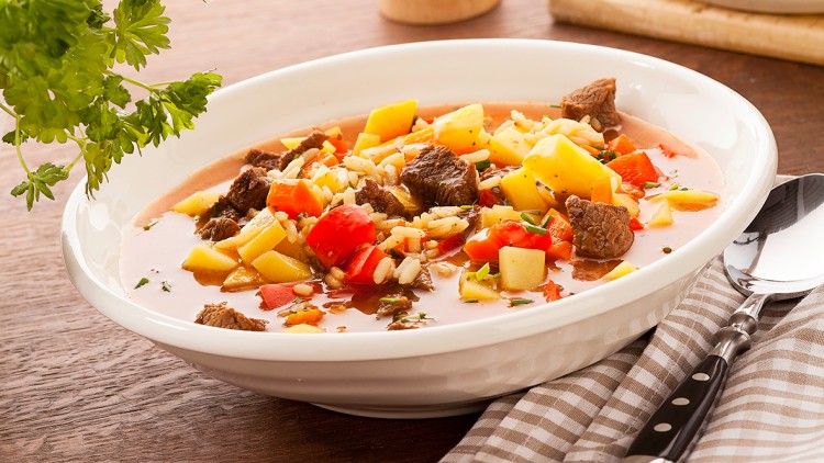 Weiße Schale mit Gemüsesuppe, Fleischstücken, Reis und Kräutern, daneben ein Löffel auf gestreiftem Stoff, im Hintergrund Petersilie.