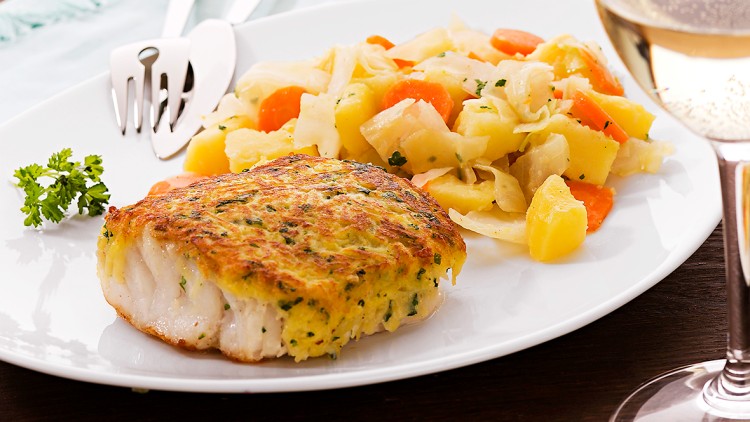 Auf weißem Teller liegen panierter Fisch, Gemüse mit Kartoffeln, daneben Besteck, ein Glas Weißwein und eine Karaffe.