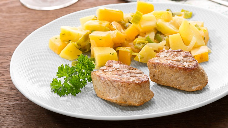Zwei gebratene Fleischmedaillons mit Petersilie und Kartoffeln auf einem weißen Teller, daneben Besteck und ein Glas Weißwein.