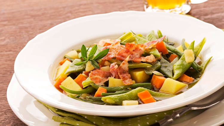Teller mit Gemüsesuppe, grünen Bohnen, Karotten, Kartoffeln und Speck, daneben ein Glas Bier auf Holztisch.