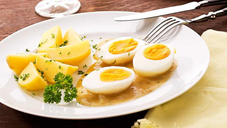 Gekochte Eier mit Senfsoße, Kartoffelstücken und Petersilie auf einem weißen Teller, daneben zwei Gläser mit hellem Getränk.