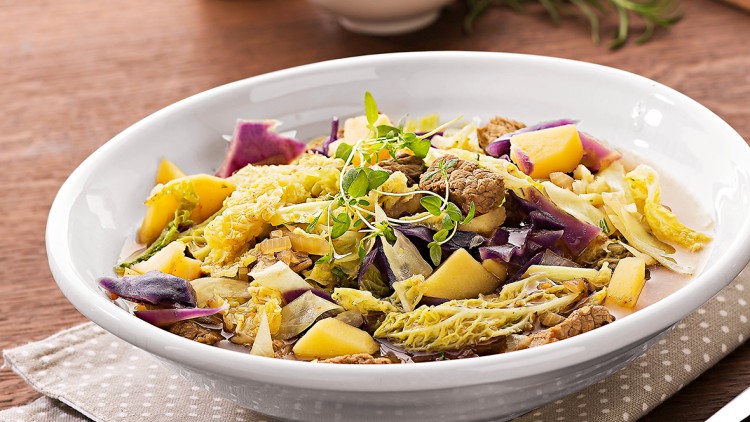Weiße Schüssel mit Eintopf aus Wirsing, Rotkohl, Kartoffeln und Kräutern, daneben Brot, Salzschale, Holzpfeffermühle und Besteck.