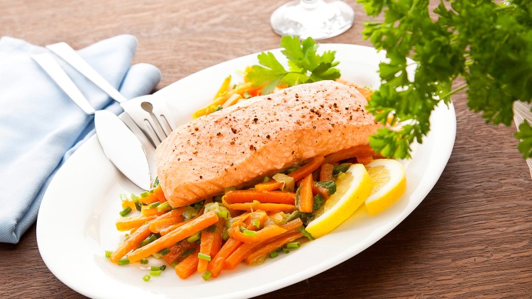 Ein Teller mit Lachsfilet auf gebratenem Gemüse, Zitronenspalten und Petersilie, daneben Besteck, ein Weinglas und ein Kräuterzweig.