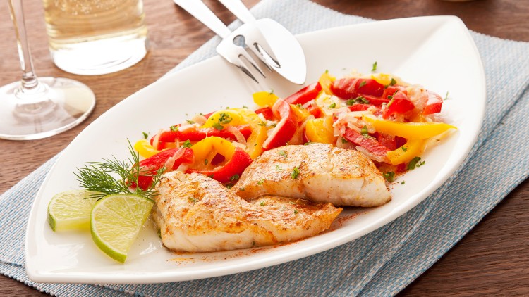 Gegrilltes Fischfilet mit Limettenscheiben, Paprika-Gemüse und Kräutern auf weißem Teller, daneben Besteck und Servietten.
