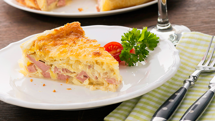 Ein Stück Quiche mit Schinken und Käse auf einem weißen Teller, dazu Deko aus Petersilie und Kirschtomate, mit Rotweinglas.
