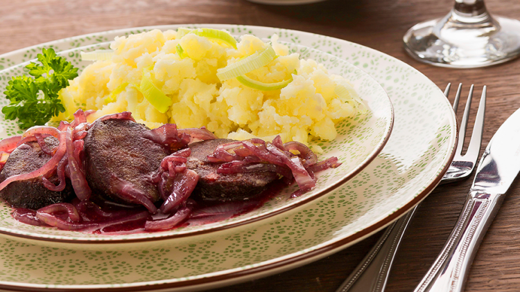 Ein Teller mit Kartoffelstampf und gebratener Wurst mit roten Zwiebeln, daneben ein Glas Rotwein und Besteck.