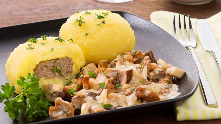 Zwei Kartoffelknödel mit Kräutern, gefüllt mit Fleisch, serviert mit Pilzsoße, Petersilie und Besteck auf einem Holztisch.