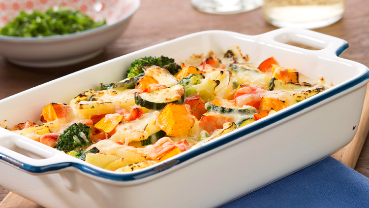 Auflaufform mit überbackenem Gemüse wie Zucchini, Paprika und Brokkoli, Holzbrett und blaue Serviette darunter.