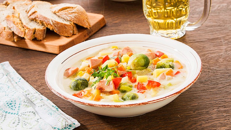 Teller mit Gemüsesuppe, Brotkorb, ein Bierkrug mit Schaum und eine Serviette auf einem Holztisch.