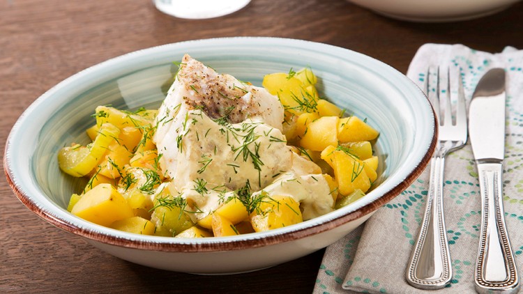 Ein Teller mit Kartoffelwürfeln, Fischfilet, heller Soße und Dill auf Holztisch, daneben Besteck auf gemusterter Stoffserviette.