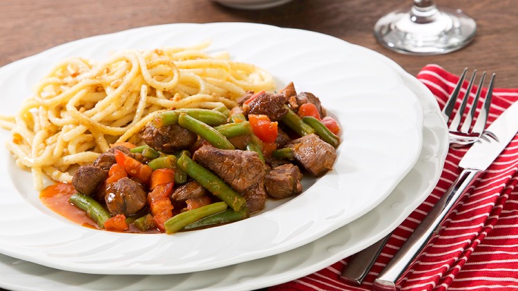 Ein Teller mit Spätzle, Rindfleisch, grünen Bohnen und Tomaten, daneben Besteck auf einer rot-weiß gestreiften Serviette.