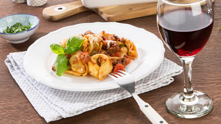 Ein Teller mit gefüllten Tortellini in Tomatensoße, garniert mit Basilikum, daneben ein Glas Rotwein und eine Auflaufform im Hintergrund.