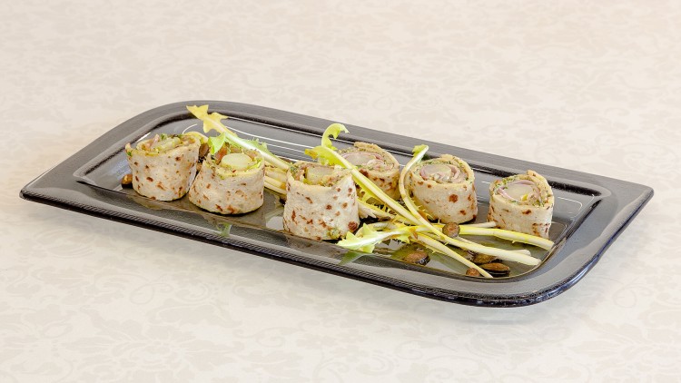 Sechs gefüllte Wrap-Röllchen mit grünem Salat und Kernen auf schwarzem rechteckigem Teller auf weißer Tischdecke.