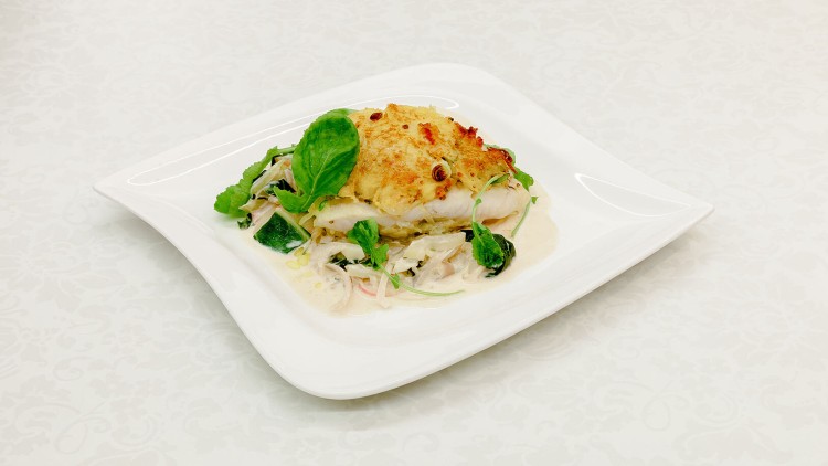 Ein weißer Teller mit gratiniertem Fischfilet auf cremigem Gemüse, dekoriert mit frischen grünen Blattspitzen.