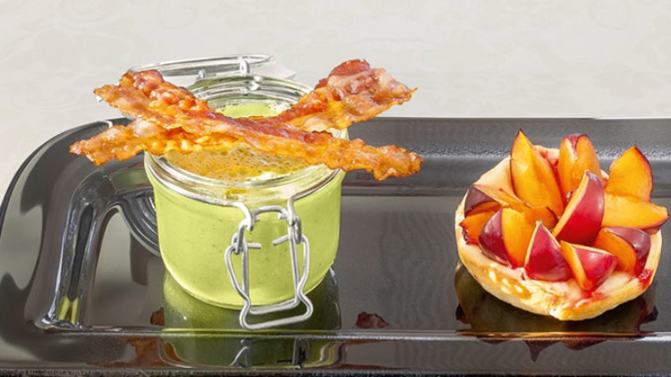 Ein Glas mit grüner Creme, darauf zwei Streifen knuspriger Speck, daneben eine Obsttarte mit Pflaumen auf schwarzem Tablett.